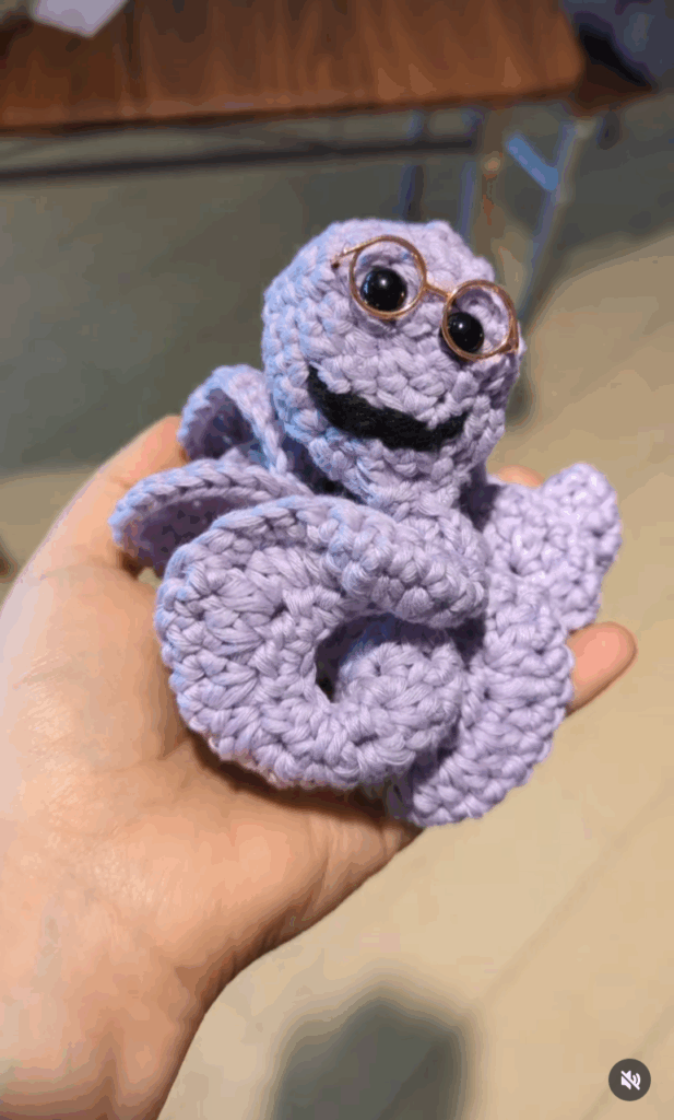 Ein gehäkeltes lilafarbenes Lesewürmchen auf der Hand.