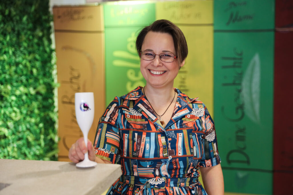 Sissi trägt ein Bücherkleid und hat ein Gals Secco in der Hand. Sie lächelt dem Betrachter einladend zu.