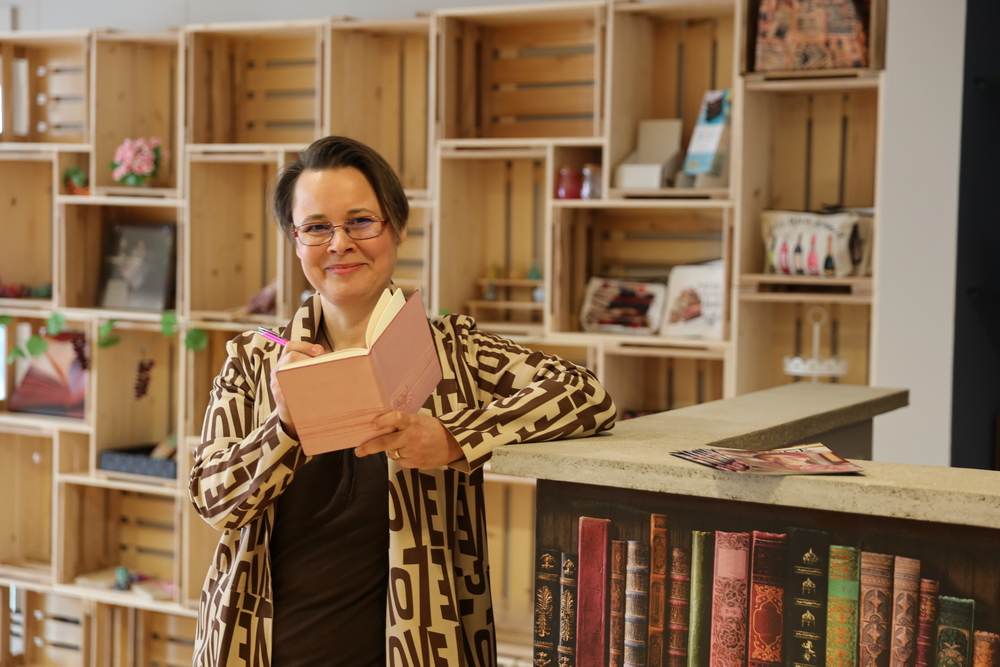 Sissi steht an ihrer Büchertheke mit einem Notizbuch in der Hand, in das sie schriebt. Sie lächelt in die Kamera.